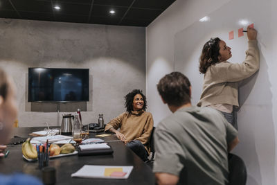 Young businesswoman writing on adhesive notes by male and female colleagues in board room