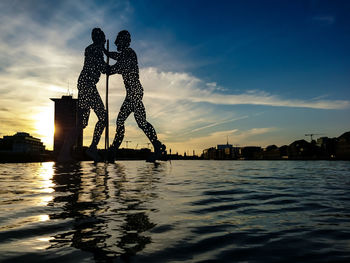 Silhouette sculpture by river against sky during sunset