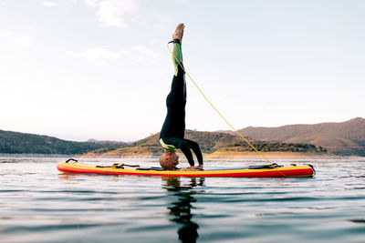 Side view of male surfer practicing yoga in headstand pose on sup board in sea at sunset
