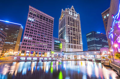 Illuminated modern buildings by river against sky at night