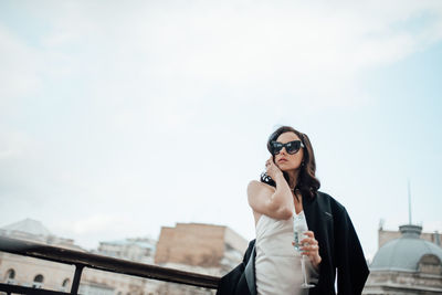 Low angle view of woman wearing sunglasses and champagne flute standing by railing against sky