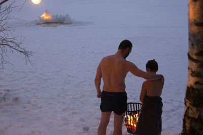 Rear view of shirtless father standing with son by bonfire at night