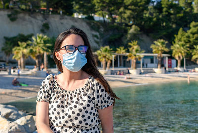 Young woman wearing surgical mask, sitting and relaxing on beach in summer.