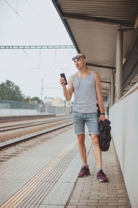 Young man in a t-shirt on the platform waiting for a train using mobile phone. man by train station