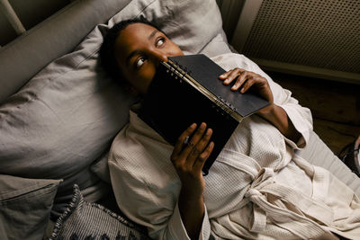 High angle view of thoughtful woman with book lying on bed at home