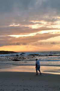 Rear view of man walking on beach against cloudy sky during sunset