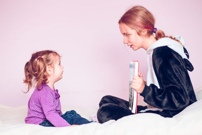 Girl showing book to sister on bed against wall at home