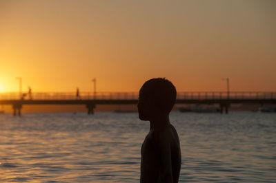 Side view of silhouette boy standing by sea against sky during sunset
