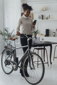 Mid adult woman leaving her home office, pushing bicycle, using smartphone