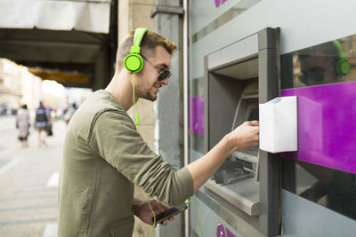 Side view of smiling man using atm and smart phone