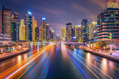 Light trails over river amidst buildings against sky at night