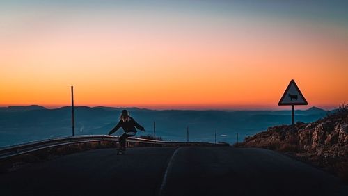 Rear view of woman skateboarding on road against clear sky during sunset