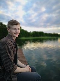 Portrait of teenage boy sitting by lake against sky