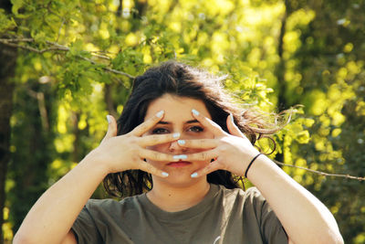 Portrait of young woman peeking through fingers against trees