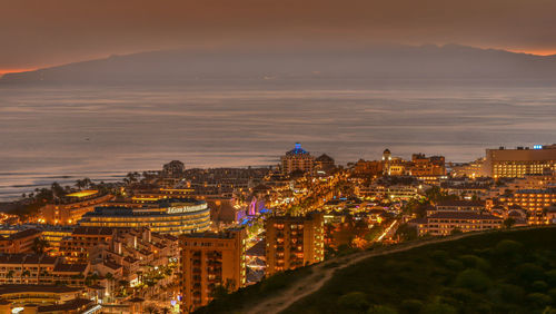 Illuminated cityscape by sea against sky at sunset