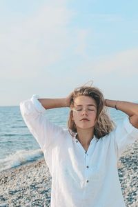 Teenage girl with eyes closed and hands in hair standing at beach against sky