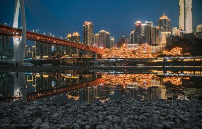 Illuminated bridge over river by buildings against sky at night