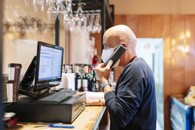 Male owner in mask taking orders through phone while standing in front of computer during pandemic