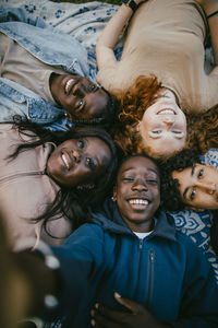 High angle view of smiling teenage boy taking selfie with male and female friends lying down on picnic blanket