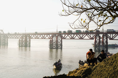 People sitting on bridge over river against clear sky