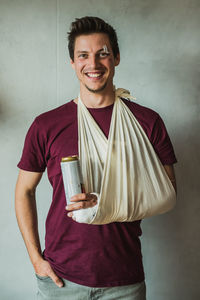 Portrait of smiling young man with fracture hand holding alcohol can while standing against wall
