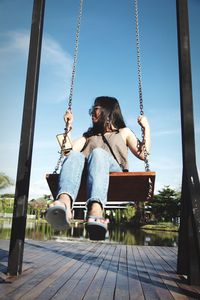 Woman sitting on swing at playground against lake and sky