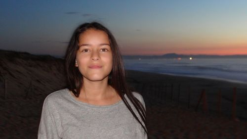 Portrait of teenage girl standing at beach against sky during sunset