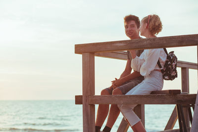 Couple looking at sea while sitting on pier against sky during sunset