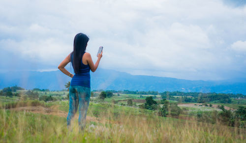 Rear view of woman using phone while standing on landscape against cloudy sky