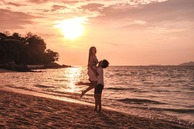 Man carrying woman while standing at beach against sky during sunset