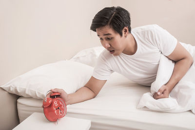 Worried young man looking at alarm clock while lying on bed