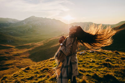 Side view of woman tossing hair while standing on mountain against sky during sunset