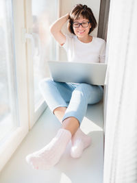 Smiling woman works remotely from home. she sits on window sill with laptop on knees.