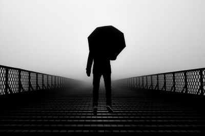 Rear view of silhouette man with umbrella levitating over bridge against clear sky