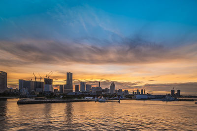 Sea by city buildings against sky during sunset