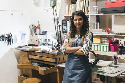 Portrait of confident female artisan with arms crossed standing against shelves in workshop