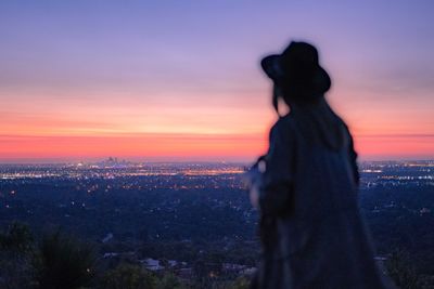 Woman looking at illuminated cityscape against sky during sunset