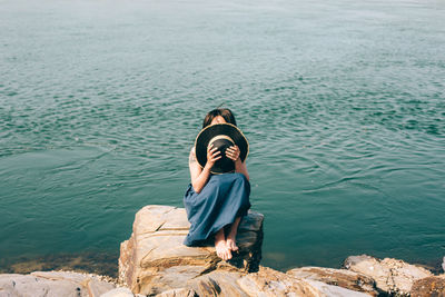 Woman covering face with hat while sitting on rock by sea