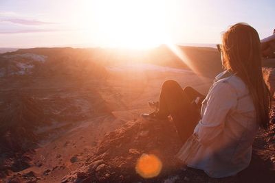 Rear view of woman sitting on cliff against sky during sunset