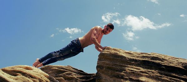 Low angle view of shirtless man exercising on rock against sky