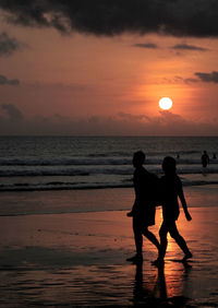 Silhouette couple walking at beach against orange sky