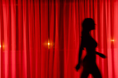 Side view silhouette of unrecognizable enigmatic skinny female walking behind red curtain near window overlooking bright streetlight at night time