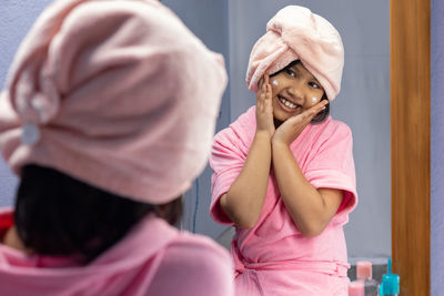 A cute indian girl child in pink bathrobe applying face cream and touching cheeks in front of mirror