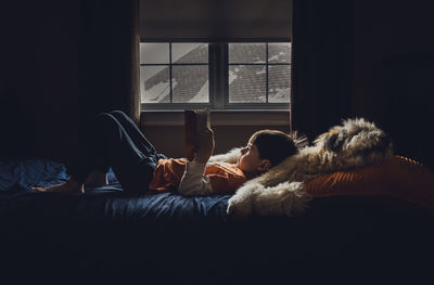 Side view of boy reading book while lying on bed at home