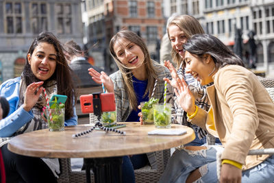 Four multi ethnic female friends sitting at a cafe terrace using mobile phone for a video call