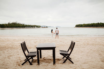 Rear view of couple at beach with table and chairs in foreground