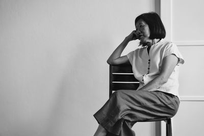 Senior woman looking away while sitting on chair against wall