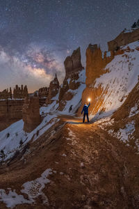 Silhouette of unrecognizable explorer standing with flashlight on scenery of rocky formations in snowy mountains under milky way starry sky in goblin valley state park, utah, usa.