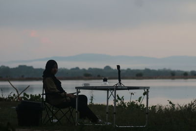 Man sitting on seat by lake against sky during sunset