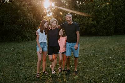 Happy adult couple with adorable daughters standing on grass near green trees and smiling while resting together and enjoying summer day in nature